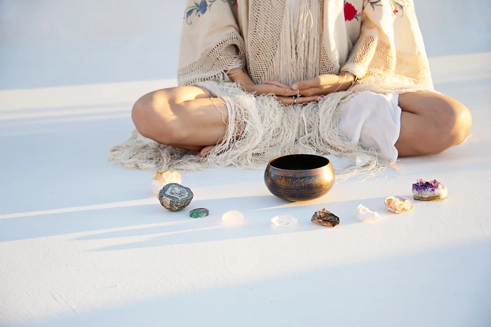 Person sitting cross-legged on a white surface with a singing bowl and crystals arranged in front, in peaceful neutral tones
